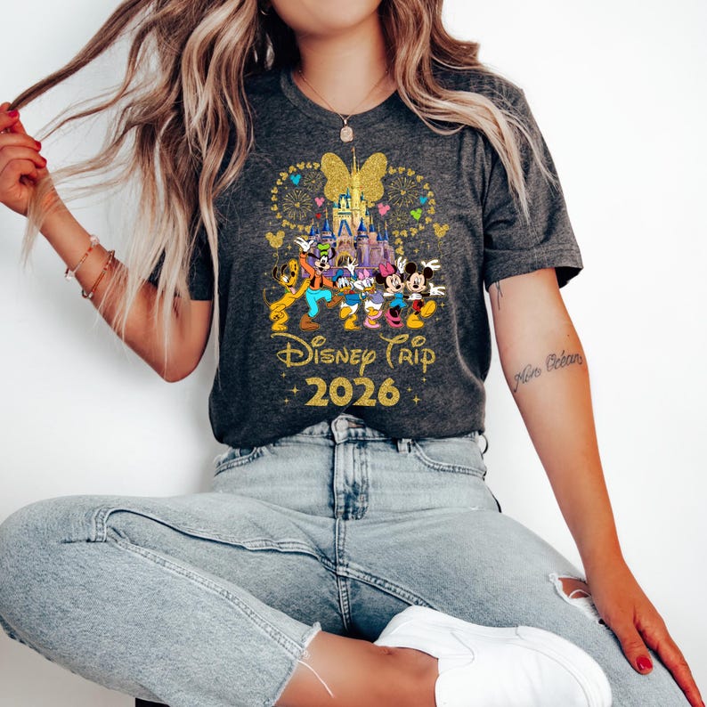 May include: Dark gray t-shirt featuring a colorful graphic of Disney characters in front of a castle, with fireworks and a butterfly design. The text "Disney Trip 2026" is in gold. The shirt is short-sleeved.