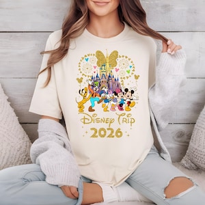 May include: Cream-colored t-shirt featuring a gold glitter design of Disney characters, a castle, and a butterfly. The text "Disney Trip 2026" is also in gold glitter. The shirt has a crew neck and short sleeves.