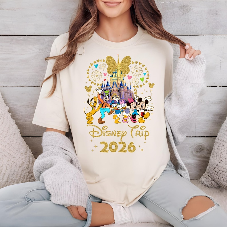 May include: Cream-colored t-shirt featuring a gold glitter design of Disney characters, a castle, and a butterfly. The text "Disney Trip 2026" is also in gold glitter. The shirt has a crew neck and short sleeves.