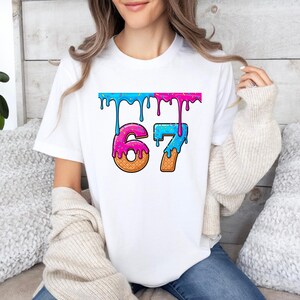 May include: White t-shirt featuring the number "67" in a playful, colorful design. The numbers are styled to resemble ice cream cones with dripping blue and pink frosting, and sprinkles. The shirt is a casual, everyday wear item.