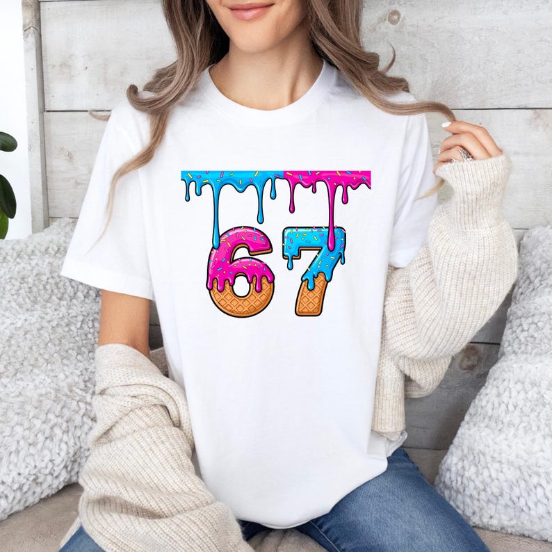 May include: White t-shirt featuring the number "67" in a playful, colorful design. The numbers are styled to resemble ice cream cones with dripping blue and pink frosting, and sprinkles. The shirt is a casual, everyday wear item.
