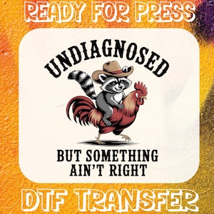 May include: DTF transfer design featuring a raccoon wearing a cowboy hat riding a rooster. The text reads "UNDIAGNOSED" above the image and "BUT SOMETHING AIN'T RIGHT" below. The words "READY FOR PRESS" and "DTF TRANSFER" are also included.