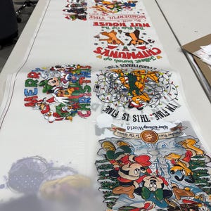 May include: A long, white sheet featuring various cartoon designs, including Chipmunks, Mickey Mouse, and holiday-themed graphics. Text includes "NUT HOUSE", "THIS IS FINE", and "WALT DISNEY World". The sheet is laid out on a white table.