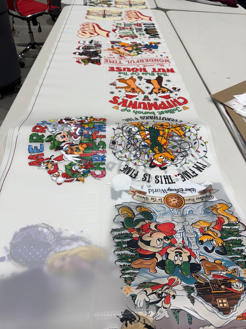 May include: A long, white sheet featuring various cartoon designs, including Chipmunks, Mickey Mouse, and holiday-themed graphics. Text includes "NUT HOUSE", "THIS IS FINE", and "WALT DISNEY World". The sheet is laid out on a white table.