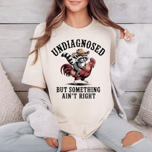 May include: Cream-colored t-shirt featuring a graphic of a raccoon wearing a cowboy hat riding a rooster. The text "UNDIAGNOSED" is arched above the image, with "BUT SOMETHING AIN'T RIGHT" below. The design is in black, red, and gray.