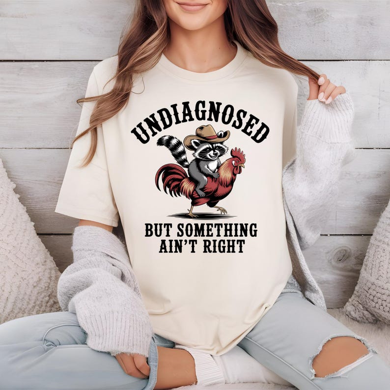May include: Cream-colored t-shirt featuring a graphic of a raccoon wearing a cowboy hat riding a rooster. The text "UNDIAGNOSED" is arched above the image, with "BUT SOMETHING AIN'T RIGHT" below. The design is in black, red, and gray.