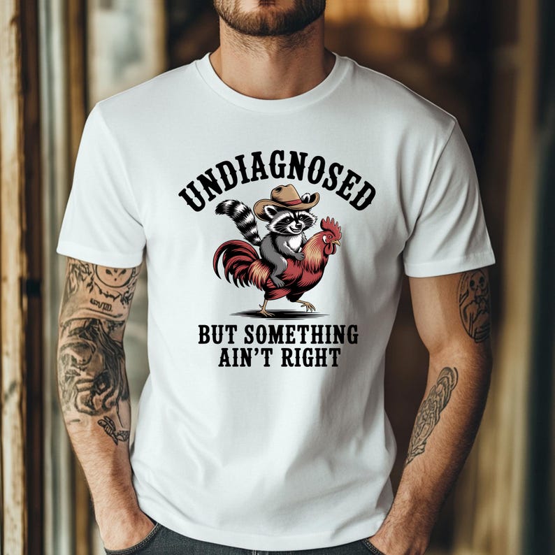 May include: White t-shirt featuring a graphic of a raccoon wearing a cowboy hat riding a red rooster. The text "UNDIAGNOSED" is above the image, and "BUT SOMETHING AIN'T RIGHT" is below. The shirt is worn by a person.