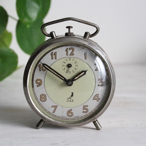 Op de afbeelding: Een ronde vintage wekker met een zilverkleurige metalen rand, een lichtgroene en witte wijzerplaat en zwarte wijzers. De klok heeft bruine Arabische cijfers, een kleine sub-wijzerplaat en een vogellogo boven de tekst JAZ en MADE IN FRANCE.