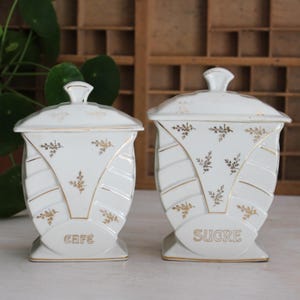 Op de afbeelding: Twee witte keramische voorraadpotten met deksels, voorzien van gouden bloemmotieven en gouden randen. De kleinere pot is gelabeld met CAFE en de grotere met SUCRE. Beide potten hebben een getrapte, geometrische vorm met een vierkante voet.