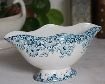 May include: A white ceramic gravy boat with a teal floral pattern. The boat has a scalloped edge and sits on a pedestal base. The floral design features daisies and leaves. A vintage style piece.