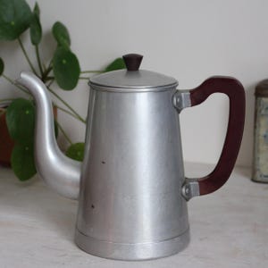 Puede incluir: Una cafetera de metal color plata con un pico curvo, una tapa a juego con un pomo marrón oscuro y un asa marrón oscuro. El cuerpo de la cafetera tiene un acabado cepillado suave, una forma ligeramente cónica y una base ancha para mayor estabilidad.
