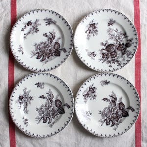 Peut inclure: Quatre assiettes en céramique blanche avec un motif floral dans les tons marron et gris. Chaque assiette présente un motif floral détaillé au centre et une bordure décorative. Les assiettes sont disposées sur une surface blanche avec des rayures rouges.