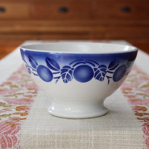 Peut inclure: Un bol en céramique blanche avec une base sur pied, orné d'une bande décorative bleue autour du bord supérieur. La bande présente un motif répétitif de formes de fruits circulaires bleues entremêlées de feuilles stylisées contrastantes.