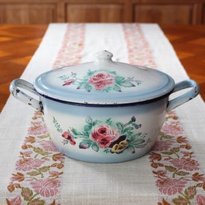 Op de afbeelding: Een witte emaille pan met deksel, voorzien van een blauwe gradiëntrand en bloemmotieven van roze rozen en groene bladeren. De pan heeft twee zijhandgrepen met zichtbare slijtage en donkere vlekken, en een knop in het midden van het deksel.