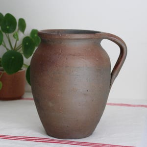 Könnte beinhalten: Ein dunkelbrauner, rustikaler Keramikkrug mit bauchigem Körper und einem einzelnen geschwungenen Henkel. Die Oberfläche des Gefäßes weist eine matte, erdige Textur mit subtilen Farbvariationen auf und zeigt ein schlichtes, funktionales Design.