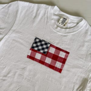 May include: White t-shirt with a red and black checkered patch sewn on the front. The patch is in the shape of a flag. The t-shirt has a tag that says "Comfort Colors" and "M-M-M".