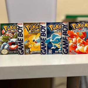 Könnte beinhalten: Fünf verschiedene Game Boy-Module für die Pokemon-Videospielserie. Die Module sind von links nach rechts angeordnet: Grün, Gelb, Blau, Rot. Die Module sind alle in ihrer Originalverpackung und zeigen das ikonische Pokemon-Logo und die Charaktere. Der Text auf den Modulen lautet "Game Boy", "Pokemon", "Gotta catch 'em all!" und die Versionsnamen.