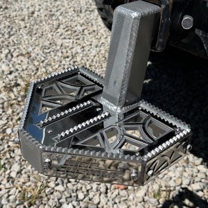 May include: A silver-colored, custom-made step with a textured, geometric design. The step is attached to a vehicle's hitch and features the text "RICE CUSTOMS". The step has a sturdy, angular construction.