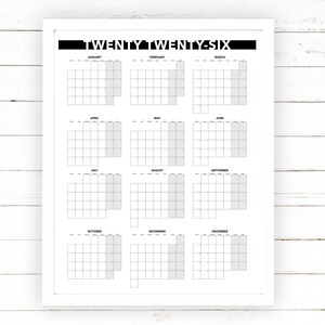 May include: A white framed calendar print for the year "TWENTY TWENTY-SIX". The calendar features a monthly layout with each month's days arranged in a grid. The days of the week are not labeled. The background is a white wooden surface.