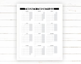 May include: A white framed calendar print for the year "TWENTY TWENTY-SIX". The calendar features a monthly layout with each month's days arranged in a grid. The days of the week are not labeled. The background is a white wooden surface.