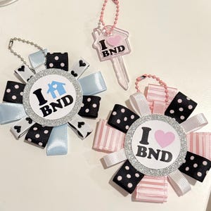 Pode incluir: Duas rosetas de fita circulares com bordas de glitter prateado e o texto I [ícone de casa] BND e I [ícone de coração] BND, acompanhadas por um pequeno chaveiro rosa translúcido em forma de chave com o texto I [ícone de coração] BND.