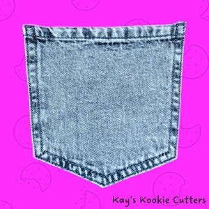 May include: A denim pocket-shaped cookie cutter with a light blue wash. The pocket has detailed stitching around the edges and a classic design. The background is bright pink with cookie-shaped designs and the text "Kay's Kookie Cutters".