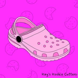 May include: A pink clog-style cookie cutter with a strap and ventilation holes. The cookie cutter is set against a pink background with a pattern of cookie outlines. The text "Kay's Kookie Cutters" is in the lower right corner.
