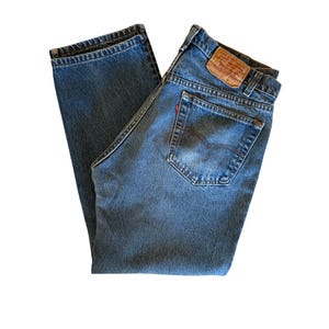 May include: Blue denim jeans with a classic five-pocket design. The jeans have a faded wash and a leather patch on the back waistband. A small red tag is visible on the back pocket. The jeans are folded.