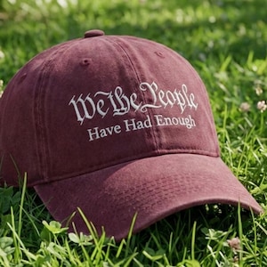 May include: A maroon baseball cap with the text "We The People Have Had Enough" in white script. The cap is resting on a bed of green grass.