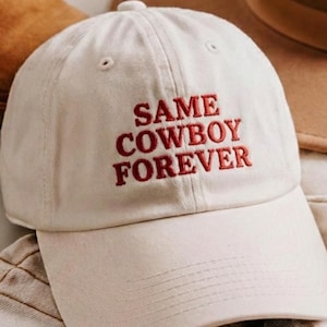 Può includere: Un cappellino da baseball color crema con la scritta ricamata in rosso "SAME COWBOY FOREVER". Il cappellino è mostrato con altri accessori, tra cui un cappello e stivali marroni.