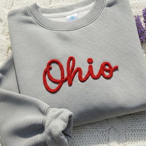 Op de afbeelding: Een grijze sweatshirt met het woord "Ohio" in het rood geborduurd. De sweatshirt ligt op een wit gebreid oppervlak, met een takje paarse lavendel rechts. De sweatshirt heeft een ronde hals en lange mouwen.