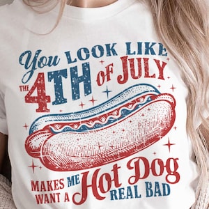 May include: White t-shirt with a graphic design featuring a hot dog and text that reads "You look like the 4th of July makes me want a hot dog real bad." The design is in red and blue.