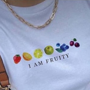 May include: White t-shirt with a graphic of a line of fruit, including a strawberry, orange, lemon, lime, apple, blueberries, and cherries. The text "I AM FRUITY" is printed below the fruit.