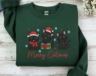 May include: A dark green sweatshirt with an embroidered design of three black cats in Santa hats and fairy lights. The words "Merry Catmas" are written in red script. The sweatshirt is folded on a light wood surface.