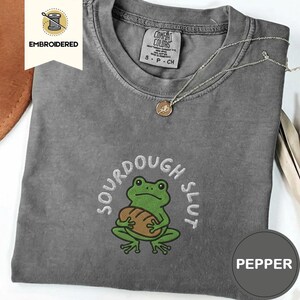 Puede incluir: Camiseta gris con un diseño bordado. Presenta una rana verde sosteniendo una hogaza de pan, con las palabras "SOURDOUGH SLUT" encima. La camiseta tiene cuello redondo y mangas cortas. Se ve un collar plateado.