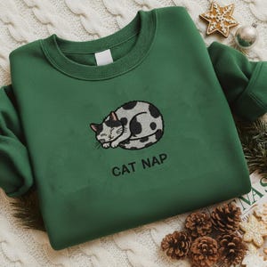 Könnte beinhalten: Ein grünes Sweatshirt mit einem gestickten Design einer schlafenden Katze mit schwarzen Flecken. Der Text "CAT NAP" befindet sich unter der Katze. Das Sweatshirt wird auf einer cremefarbenen Strickoberfläche präsentiert.