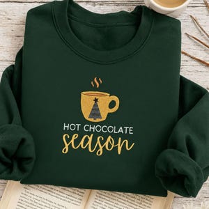 Peut inclure: Un sweat-shirt vert foncé avec l'inscription "HOT CHOCOLATE SEASON" en lettres dorées. Le motif représente une tasse dorée de chocolat chaud avec un sapin de Noël à l'intérieur. C'est un vêtement confortable à manches longues.