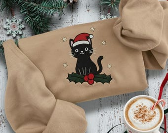 May include: A tan sweatshirt with an embroidered black cat wearing a Santa hat, surrounded by stars and holly. A cup of coffee with a candy cane is nearby. The sweatshirt is folded, with the hood visible. Christmas-themed.