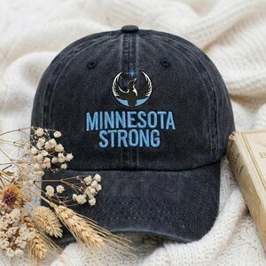 May include: A dark blue baseball cap with the embroidered text "MINNESOTA STRONG" in light blue. The cap features a circular emblem with a bird design. The hat is made of a soft, worn material.