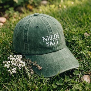 May include: A green baseball cap with the text "NEEDS SALT" embroidered in white. The cap is resting on a bed of green grass, with small white flowers and sprigs of herbs nearby. The cap has a curved brim.