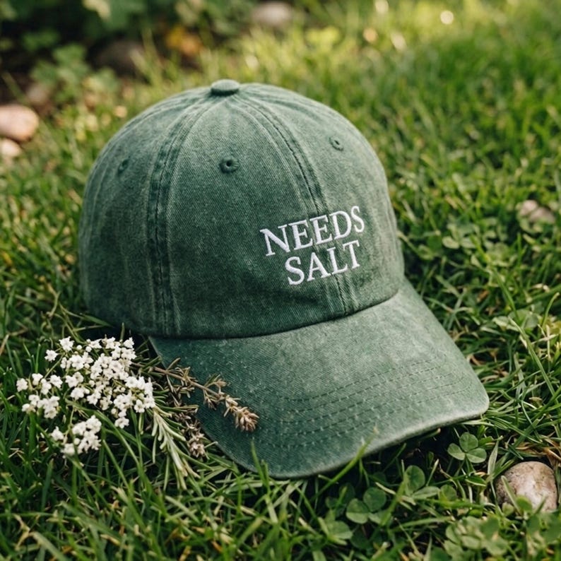 May include: A green baseball cap with the text "NEEDS SALT" embroidered in white. The cap is resting on a bed of green grass, with small white flowers and sprigs of herbs nearby. The cap has a curved brim.