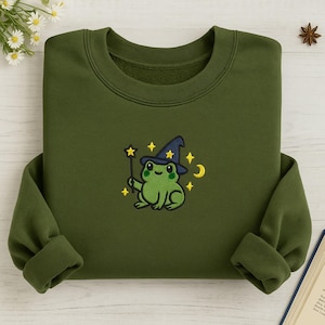 Puede incluir: Sudadera verde oliva con un diseño bordado de una rana verde con un sombrero de mago azul y una varita de estrella. El diseño incluye estrellas amarillas y una luna creciente. La sudadera tiene cuello redondo y mangas largas.