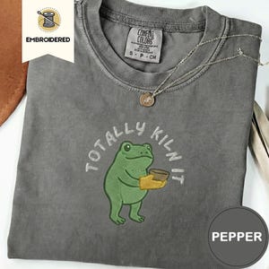 May include: Pepper-colored t-shirt with an embroidered frog holding a bowl, and the text "TOTALLY KILN IT". The shirt has a Comfort Colors tag and a silver necklace. The word "EMBROIDERED" is on a white tag.