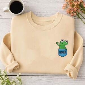 May include: A cream-coloured sweatshirt with a cartoon lizard waving from a blue pocket. The word "LIZARD" is printed on the pocket. A cup of coffee and flowers are in the background.