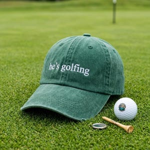 Könnte beinhalten: Eine grüne Baseballkappe mit dem weißen Text "he's golfing". Die Kappe liegt auf einem Golfplatz, neben einem Golfball, einem Tee und einem Ballmarker.