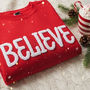 Może przedstawiać: Złożona czerwona bluza z napisem "BELIEVE" w białych, teksturowanych literach. Bluza leży na białej powierzchni z warkoczowym splotem, z kubkiem bitej śmietany i laską cukrową w tle. Szyszki i zieleń dopełniają świąteczny motyw.