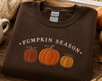 May include: A dark brown crewneck sweatshirt featuring an embroidered design of three pumpkins in varying shades of orange and the text PUMPKIN SEASON arched above them. The garment has a Gildan brand label on the inside collar indicating size M.
