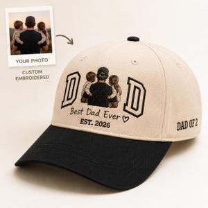 May include: A beige and black baseball cap with the word "DAD" embroidered in black, along with a graphic of a father and two children. The text "Best Dad Ever" and "EST. 2026" are also embroidered on the cap. The words "DAD OF 2" are on the side.