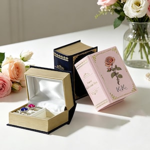 May include: A gold and navy blue book-shaped jewellery box, open to reveal two rings with blue and pink gemstones. A pink book-shaped box with a rose illustration and the text "Faerie Ballerina Fox" is also visible. A bouquet of roses sits nearby.