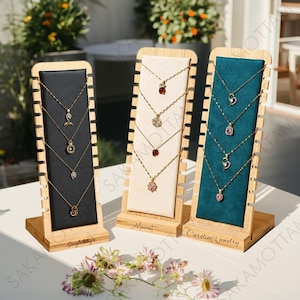 以下が含まれることがあります： さまざまなペンダントデザインのゴールドネックレスを展示する3つの木製ジュエリーディスプレイ。ディスプレイは、黒、クリーム、ティールブルーの背景です。最初のディスプレイには「Singletary's」、2番目には「Mariet」、3番目には「Caroline's Jewelry」と書かれています。